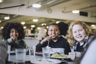 Smiling pupils looking away while sitting at table during lunch break in cafeteria