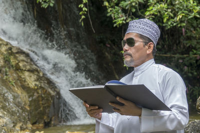 Man reading book while standing against waterfall