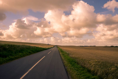 Road passing through grassy field against cloudy sky