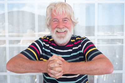 Portrait of bearded senior man holding his hands while standing against window on balcony
