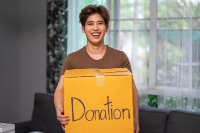 Portrait of smiling young woman holding box while standing against wall