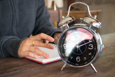 Close-up of man writing in book with alarm clock on table