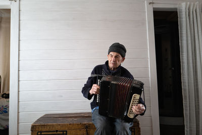 Portrait of old man playing harmonica while sitting against wall