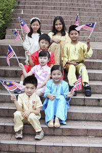 Portrait of cute friends in traditional clothing holding malaysia flags while sitting on steps