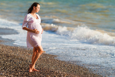 Pregnant woman stands on sea beach in summer in white light dress with fluffy
