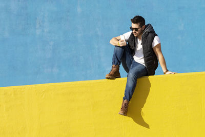 Young bearded man with sunglasses sitting on yellow wall, using phone