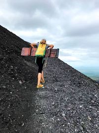 Woman carrying plank and backpack while climbing mountain against sky