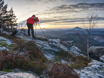 Photographer in red jacket and knitted cap stay with camera on tripod on cliff and thinking. dreamy