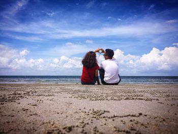 Couple making heart shape with hands while sitting at shore against cloudy sky