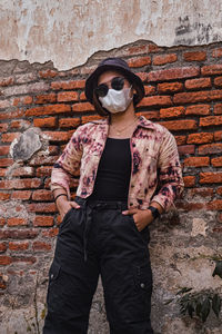 Portrait of woman wearing sunglasses and mask while standing against brick wall