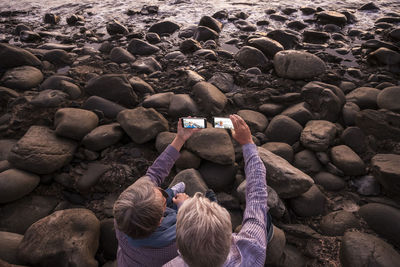 High angle view of couple taking selfie from mobile phones on rock at beach