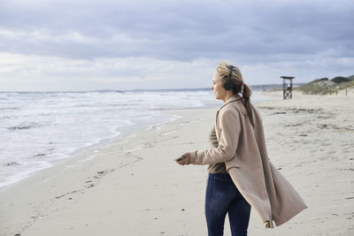 Spain, menorca, senior woman using smartphone and wireless headphones on the beach in winter