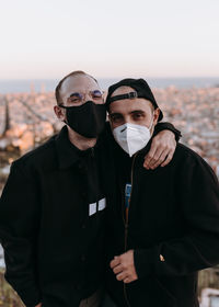 Portrait of two young men embracing and wearing face masks standing against cityscape