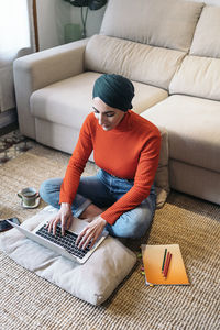 From above muslim woman in jeans sitting cross legged on cotton mat and typing on laptop keyboard while working on freelance project at home