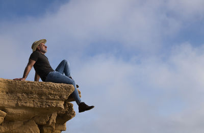 Adult man in cowboy hat sitting on cliff against sky. almeria, spain