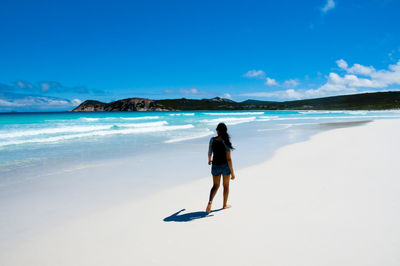 Full length rear view of woman walking at beach against blue sky on sunny day