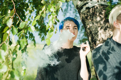 Young woman smoking electronic cigarette by tree