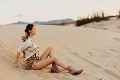 Young woman sitting on sand at beach against sky during sunset