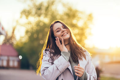 Smiling beautiful girl with long hair in a grey trench coat using smartphone in the spring 