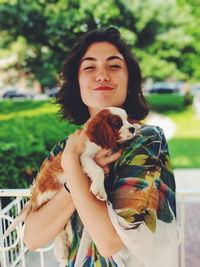 Portrait of beautiful young woman carrying puppy standing against plants in park