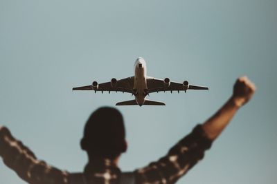 Rear view of man with arms outstretched against airplane flying in sky