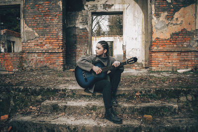 Full length of young fashionable woman with guitar while sitting on steps against abandoned building