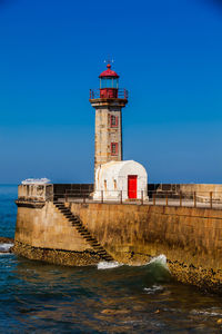 Lighthouse by sea against buildings against clear blue sky