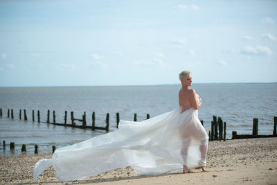 Side view of young woman with scarf standing at beach against sky