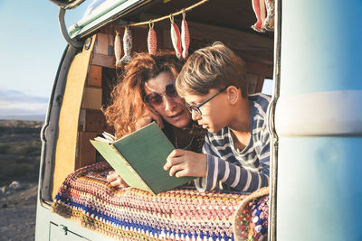 Smiling mother and son reading book while lying camper van