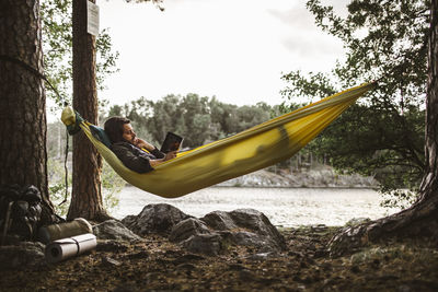 Young man reading book while lying down over hammock in forest