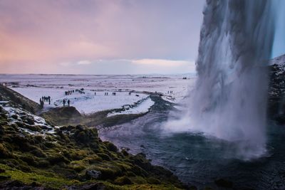 Scenic view of waterfall by snow covered landscape against cloudy sky during sunset