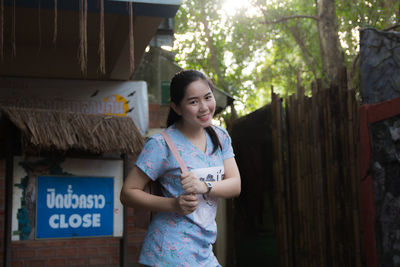 Portrait of smiling young woman standing against buildings and trees in city