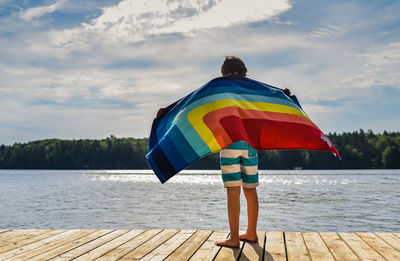 Child standing on a dock holding a rainbow towel in the wind.