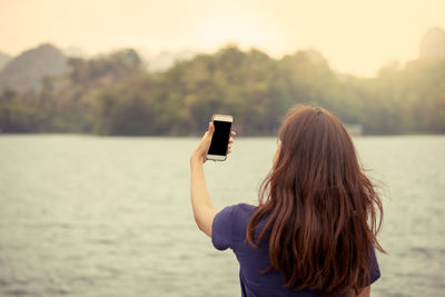 Rear view of woman using mobile phone standing against lake