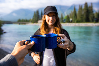 Portrait of young woman holding coffee cup against river