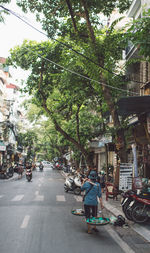 Rear view of man carrying weight scale while walking on road amidst buildings