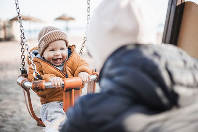 Mother pushing her cheerful infant baby boy child on a swing on sandy beach playground outdoors on