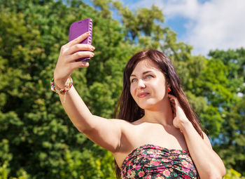 Low angle view of young woman taking selfie while smart phone while standing against trees in park