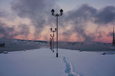 Lamp posts on snow covered field against sky during sunset