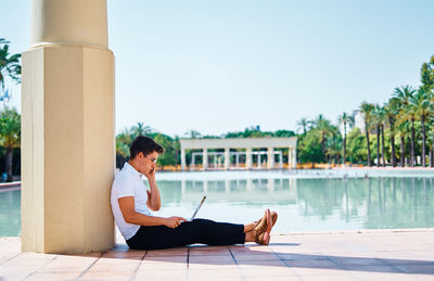 Side view of concentrated male student browsing netbook and speaking on mobile phone while sitting near stone column in university campus