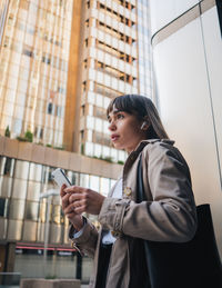 Side view of serious female in stylish outfit surfing cellphone while listening to music in wireless earphones on street with modern building