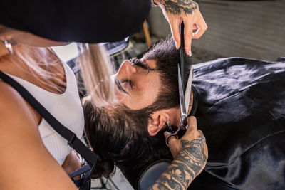 High angle of crop unrecognizable tattooed barber with scissors and comb cutting beard of stylish man sitting in chair in modern salon