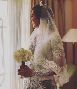 Smiling bride looking away while holding flower bouquet by window