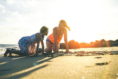 People kneeling at beach against sky during sunset