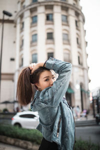 Portrait of young woman tying hair while standing on street in city