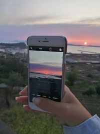 Close-up of hand holding smart phone against sunset sky