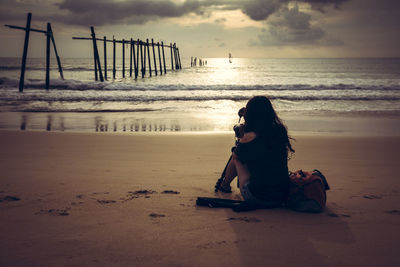 Man sitting on shore at beach against sky during sunset