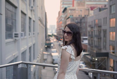 Portrait of woman wearing sunglasses while standing at balcony in city during sunset