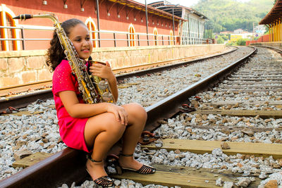 Smiling girl holding saxophone sitting on railroad track at station