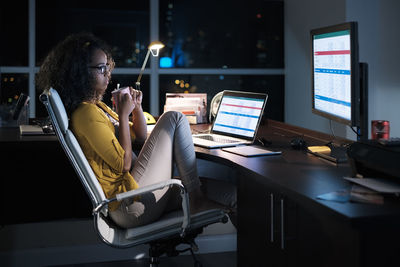 Side view of thoughtful businesswoman holding coffee cup while sitting on chair in office at night